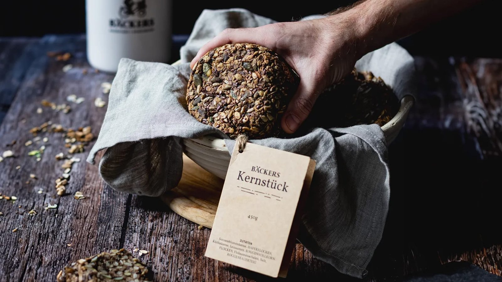 Food-Fotografie in Wien – Hand legt frisches Bäckers Kernstück mit Körnerkruste in eine rustikale Schale, stilvoll inszeniert auf Holzhintergrund.